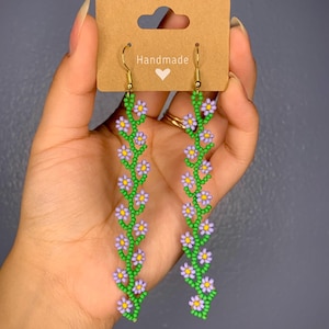 May include: A pair of dangle earrings with a floral daisy design. They are shaped like a vine going up with flowers in between. The earrings are made in multiple color ways, color combos, including: green and purple, black and white, green and blue, green and pink, green and yellow, brown and cream, green and red. The earrings are hanging from gold hooks. This beadwork is indigenous native american made