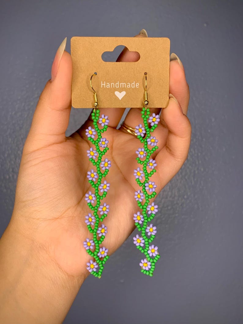 May include: A pair of dangle earrings with a floral daisy design. They are shaped like a vine going up with flowers in between. The earrings are made in multiple color ways, color combos, including: green and purple, black and white, green and blue, green and pink, green and yellow, brown and cream, green and red. The earrings are hanging from gold hooks. This beadwork is indigenous native american made