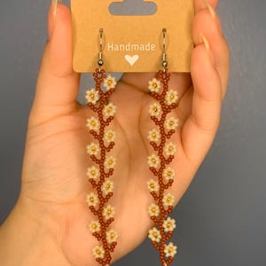 May include: A pair of long, brown beaded earrings with white flower accents. The earrings are handmade and feature a delicate floral design.