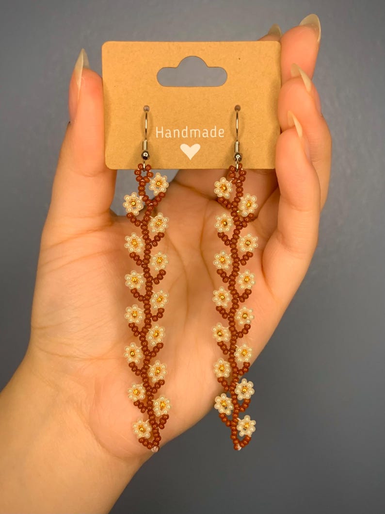 May include: A pair of long, brown beaded earrings with white flower accents. The earrings are handmade and feature a delicate floral design.