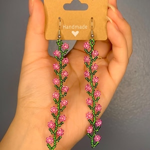 May include: A pair of handmade dangle earrings featuring pink and green beaded flowers. The earrings are on a brown card with the word "Handmade" written on it.
