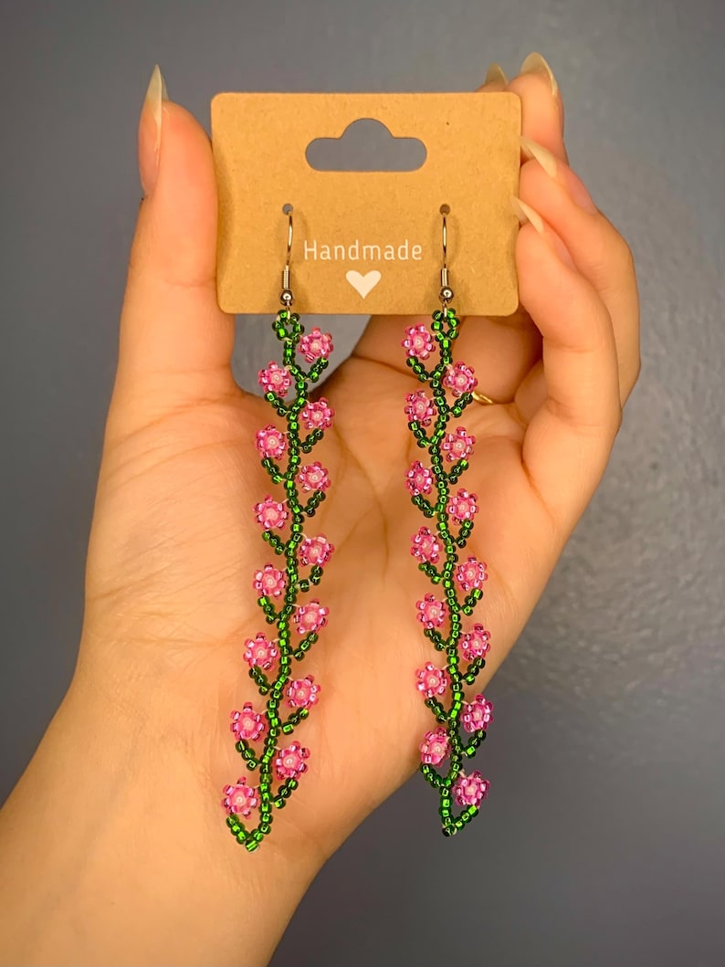 May include: A pair of handmade dangle earrings featuring pink and green beaded flowers. The earrings are on a brown card with the word "Handmade" written on it.