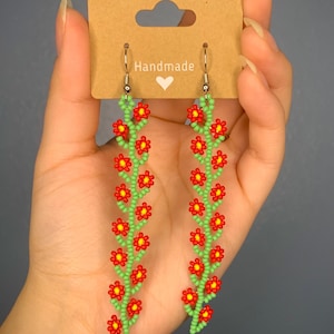 May include: A pair of handmade beaded earrings featuring a floral design. The earrings are made with red and green beads and have silver hooks.