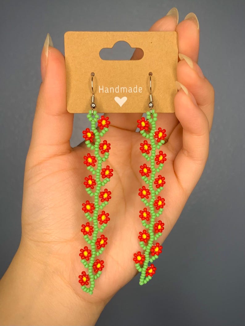 May include: A pair of handmade beaded earrings featuring a floral design. The earrings are made with red and green beads and have silver hooks.
