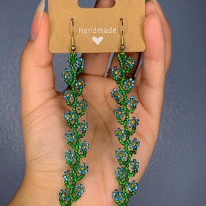 May include: Handmade beaded earrings with a floral design. The earrings are green and blue with gold accents. The earrings are hanging from gold hooks.