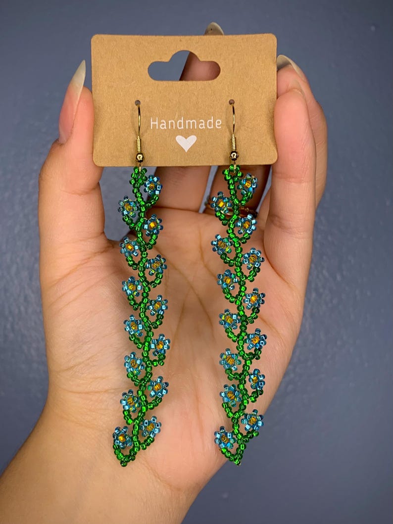 May include: Handmade beaded earrings with a floral design. The earrings are green and blue with gold accents. The earrings are hanging from gold hooks.