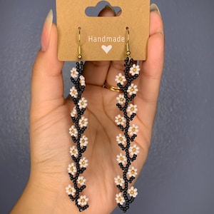 May include: A pair of handmade dangle earrings featuring a black beaded stem with white daisy-shaped beads. The earrings are on a brown card with the word "Handmade" and a heart.