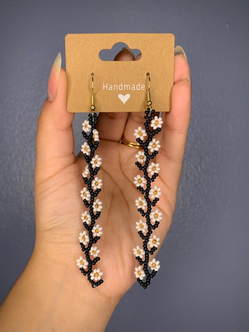 May include: A pair of handmade dangle earrings featuring a black beaded stem with white daisy-shaped beads. The earrings are on a brown card with the word "Handmade" and a heart.