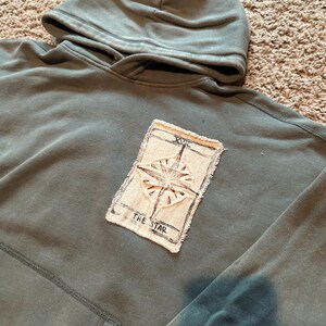 May include: A sage green hooded sweatshirt featuring a rectangular fabric patch sewn onto the center chest. The patch displays a black ink illustration of a star with the Roman numeral XVII and the text THE STAR printed below the design.