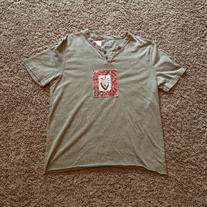 Peut inclure: Un t-shirt henley vert olive à manches courtes avec un petit écusson rectangulaire sur la poitrine. L'écusson présente une bordure à motifs rouges et blancs avec une illustration centrale en noir et blanc d'un cœur. Patte à deux boutons.