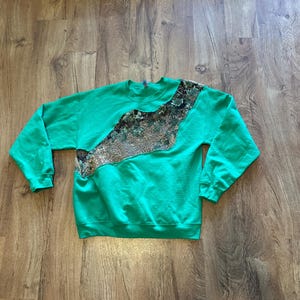 Puede incluir: Una sudadera verde con un diseño diagonal estampado en el pecho. El estampado presenta una mezcla de colores marrón, beige y gris. La sudadera tiene mangas largas y cuello redondo.