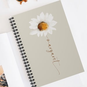 May include: A spiral bound notebook with a light green cover featuring a white daisy with a yellow center. The word "margaret" is written in cursive in brown lettering below the flower.