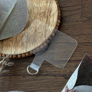 May include: A transparent, square acrylic tag with a small loop for a keyring. A wooden bead bracelet is partially visible through the tag. The background includes wooden textures and decorative leaves.