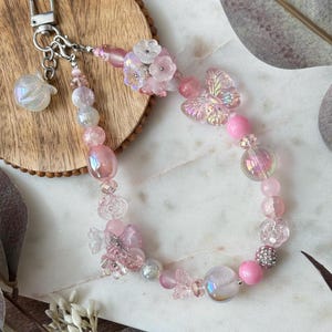 May include: A pink beaded keychain with a silver clasp and a small, iridescent apple charm. The keychain features various pink beads, including flowers and butterfly-shaped accents. The beads are strung together with a silver chain.