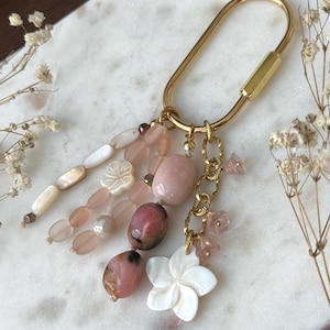 May include: A gold-toned metal oval carabiner keychain adorned with a cluster of dangling charms, including smooth pink and brown stone beads, white carved flower-shaped shell beads, and small translucent pink glass flower accents on a gold chain.