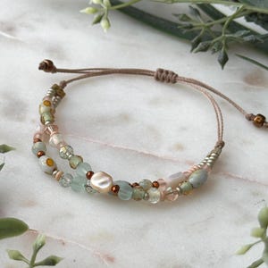 以下が含まれることがあります： パステルカラーのビーズを組み合わせた繊細なブレスレット。ピンク、グリーン、イリデセントなアクセントが含まれています。ブレスレットは、調節可能なノットクロージャーが付いた茶色のコードに通されています。デザインには、中央に真珠のようなビーズが含まれています。