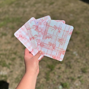 Puede incluir: Un conjunto de cuatro tarjetas de color rosa y blanco con un acabado brillante. Las tarjetas presentan un patrón de cuadros con lazos rosas.