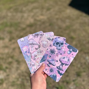 Könnte beinhalten: Vier rosa und violette Karten mit verschiedenen Designs, darunter Pilze, Schädel, Sterne und Fledermäuse.