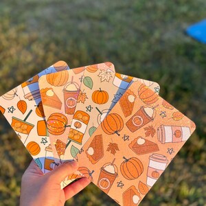 Puede incluir: Cuatro posavasos cuadrados decorativos con diseños de temática otoñal. Cada posavasos presenta un patrón diferente de calabazas, tazas de café, porciones de pastel y hojas de otoño en tonos naranja, marrón y crema. Los posavasos se sostienen en una mano.