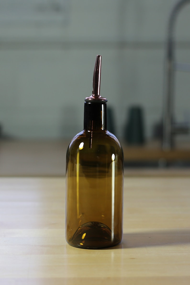 May include: A brown glass bottle with a copper pour spout. The bottle is sitting on a light wood surface.
