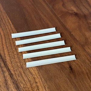 May include: Five white elastic bands, also known as ear loops, are arranged in a row on a wooden surface. The bands are all the same size and are made of a flexible material.