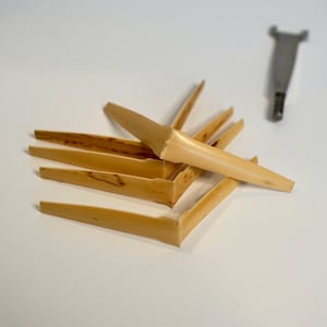 May include: A set of six beige wooden reeds for a musical instrument. The reeds are stacked on top of each other and are slightly curved. A small metal tool is visible in the background.