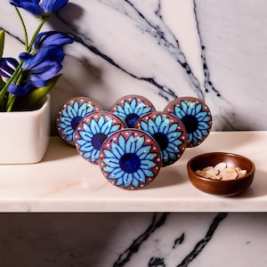 May include: Six blue and brown ceramic knobs with a floral design. The knobs have a raised, shiny blue center with a dark blue circle in the middle. The knobs are surrounded by a brown ring with a white border.