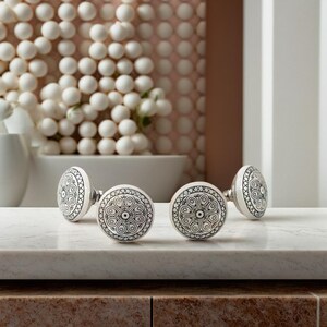 May include: Four white ceramic door knobs with intricate black and silver designs. The knobs are arranged in a row on a white marble surface.