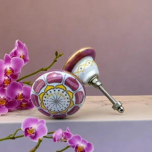 Könnte beinhalten: Zwei Keramik-Schrankknöpfe mit einer rosa-weißen Basis und einem gelben und goldenen Blumendesign. Die Knöpfe haben eine silberne Metallschraube.