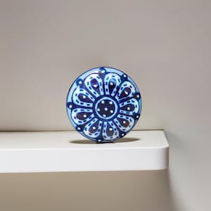 Op de afbeelding: Een ronde, blauw-witte keramische kastknop met een bloemmotief. De knop heeft een donkerblauw midden met witte stippen en een herhalend patroon van donkerblauwe bloemblaadjes. De knop ligt op een witte plank.