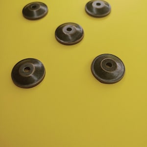 Op de afbeelding: Vijf ronde, donkerbruine metalen meubelsteunen met een verhoogde rand. De steunen zijn in een patroon op een geel oppervlak gerangschikt.