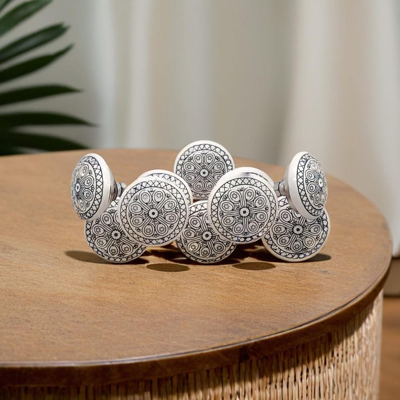 May include: A set of seven white ceramic drawer knobs with black intricate designs. The knobs are arranged in a row on a wooden surface.
