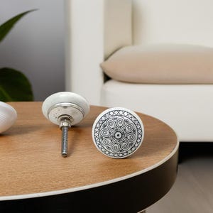 May include: Three white ceramic cabinet knobs with a silver metal base and a black and white geometric design. The knobs are on a wooden surface.
