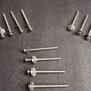 Puede incluir: Una colección de tornillos plateados con cabezas decorativas. Algunos tornillos tienen una cabeza redonda con borde estriado, otros una cabeza plana en forma de cruz. Los tornillos están dispuestos sobre una superficie gris oscuro. Medidas métricas.