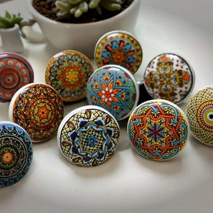 Könnte beinhalten: Neun Keramik-Schrankknöpfe mit komplizierten, bunten Mandala-Designs. Die Knöpfe sind rund und haben einen weißen Hintergrund.