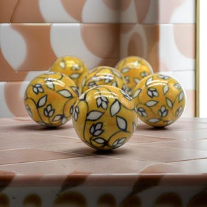 Peut inclure: Six sphères en céramique jaune avec un motif floral blanc. Les sphères sont disposées en cercle sur une surface carrelée rose et blanche.