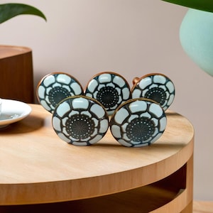 Op de afbeelding: Set van zes witte keramische ladegrepen met een geometrisch patroon in zwart en wit. De knoppen hebben een koperen afwerking rond de randen.