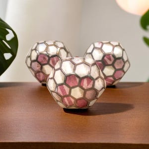 May include: Three decorative spherical objects with a geometric pattern of white and burgundy hexagons. The objects are arranged on a dark brown wooden surface. The background includes a green plant and a soft, out-of-focus light.