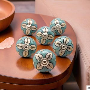 Op de afbeelding: Zes keramische kastknoppen met een blauw en wit bloemenmotief. De knoppen hebben een zilveren midden en zijn gerangschikt op een houten oppervlak.