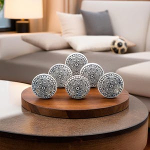 May include: Six white ceramic drawer knobs with a black and white geometric pattern. The knobs are arranged in a circle on a wooden surface.