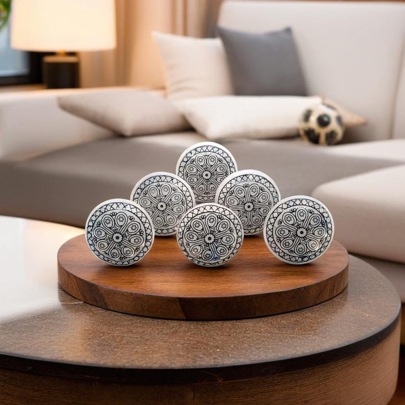May include: Six white ceramic drawer knobs with a black and white geometric pattern. The knobs are arranged in a circle on a wooden surface.