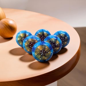 Op de afbeelding: Zes blauwe en gouden keramische knoppen met een wit en zwart abstract ontwerp. De knoppen zijn in een cirkel op een houten oppervlak gerangschikt.