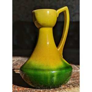 May include: A vintage ceramic pitcher with a yellow and green color scheme. The pitcher has a bulbous base, a slender neck, and a handle. The glaze has a crackle effect, adding to its antique charm. The pitcher is likely used for serving beverages.