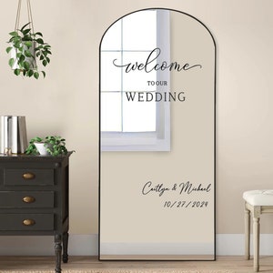 May include: A tall, arched mirror with a black frame. The mirror has a welcome sign that says "Welcome to our Wedding" and the names "Caitlynn & Michael" and the date "10/27/2024".