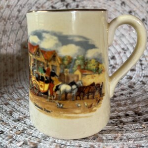 Puede incluir: Una taza de cerámica de color crema con un borde dorado y un asa. La taza presenta una ilustración vintage de un carruaje tirado por caballos y una escena de pueblo. El texto "Dicken's Days" está impreso en la parte inferior de la taza.