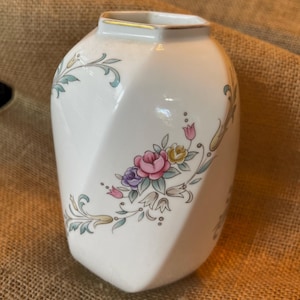 May include: A white ceramic vase with a geometric shape and floral design. The vase has a gold trim around the top and features a delicate floral pattern in shades of pink, purple, yellow, and green.