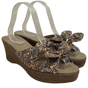 May include: A pair of wedge sandals with a floral pattern. The sandals have a brown woven sole and a strap with a large bow. The floral pattern includes brown, orange, and purple flowers on a dark background. The sandals are displayed on a white background.