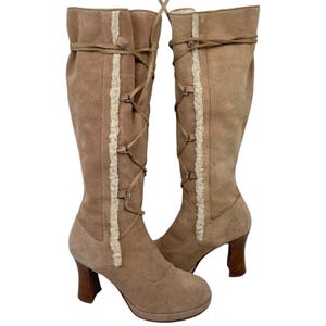 May include: A pair of knee-high tan suede boots featuring a lace-up front design with metal eyelets and a vertical strip of cream-colored faux shearling trim. The boots have a rounded toe, a platform sole, and a sturdy brown block heel.