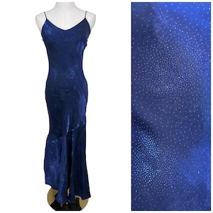 Puede incluir: Un vestido largo azul marino con tirantes finos y cuello en V. El vestido tiene un corpiño ajustado y una falda acampanada con dobladillo con volantes. La tela tiene un brillo sutil. El vestido se exhibe en un maniquí.