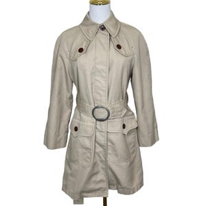 May include: A tan trench coat with a belted waist and a large silver-colored buckle. The coat has two front pockets, brown buttons, and a collared neckline. The sleeves are three-quarter length.
